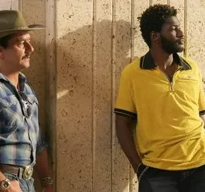 Um homem de chapéu e camisa xadrez e outro de camisa polo amarela, ambos apoiados, olhando à direita em ambiente interno.