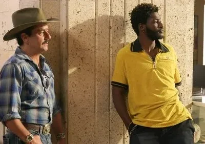 Um homem de chapéu e camisa xadrez e outro de camisa polo amarela, ambos apoiados, olhando à direita em ambiente interno.