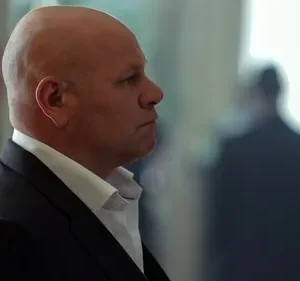 Homem careca em terno escuro com expressão séria, olhando para a direita. Fundo desfocado com silhuetas e luz forte.