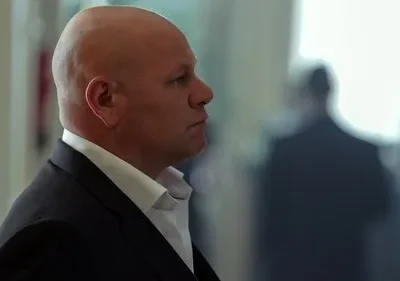 Homem careca em terno escuro com expressão séria, olhando para a direita. Fundo desfocado com silhuetas e luz forte.