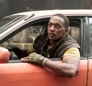 Homem negro concentrado em carro laranja desgastado. Veste colete e luvas, espreita pela janela do motorista com o braço para fora.