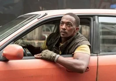 Homem negro concentrado em carro laranja desgastado. Veste colete e luvas, espreita pela janela do motorista com o braço para fora.