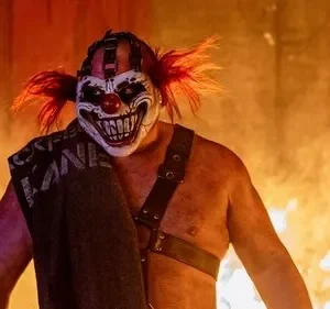 Palhaço aterrorizante de máscara branca, cabelo vermelho e sorriso dentuço, sem camisa, contra um fundo de chamas laranjas e fumaça.