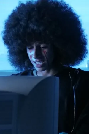 Pessoa de cabelo afro com manchas vermelhas no rosto chora intensamente, olhando para um monitor. Fundo escuro com luz azul. Clima angustiante.