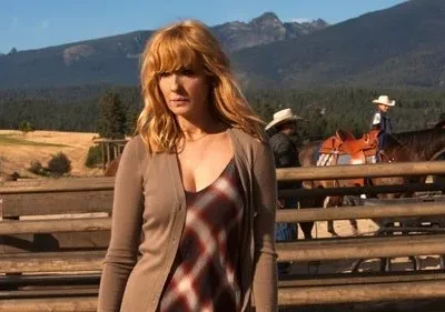 Mulher séria de cabelo loiro em cardigan, num rancho com montanhas e floresta. Pessoas a cavalo ao fundo. Atmosfera ocidental.