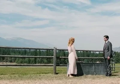 Mulher de vestido claro e homem de terno se olham em campo vasto. Cerca de madeira, floresta e montanhas ao fundo. Clima sério e bucólico.