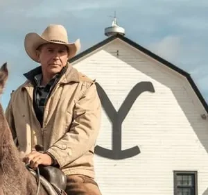 Homem sério de chapéu cowboy e jaqueta em um cavalo marrom. Ao fundo, um celeiro branco com grande 'Y' e colinas secas.
