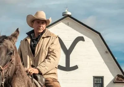 Homem sério de chapéu cowboy e jaqueta em um cavalo marrom. Ao fundo, um celeiro branco com grande 'Y' e colinas secas.