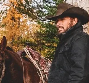 Um homem barbudo de chapéu de cowboy e jaqueta escura está ao lado de um cavalo marrom, em uma floresta de outono.