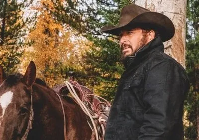 Um homem barbudo de chapéu de cowboy e jaqueta escura está ao lado de um cavalo marrom, em uma floresta de outono.