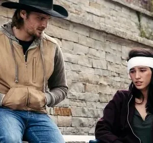 Cowboy e mulher com atadura na cabeça sentados ao ar livre, contra uma parede de pedra. Ele a observa, clima sério e preocupado.