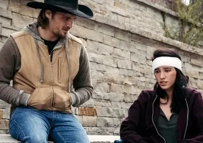 Cowboy e mulher com atadura na cabeça sentados ao ar livre, contra uma parede de pedra. Ele a observa, clima sério e preocupado.