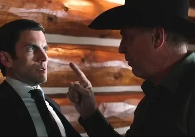 Dois homens, um de terno e outro de chapéu de caubói, em discussão tensa contra parede de troncos.