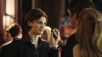 Uma mulher de cabelo curto e blazer escuro sorri, segurando um distintivo. Ela interage com um homem de costas em um ambiente noturno e movimentado.
