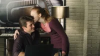 Mulher ruiva beija a testa de homem sorridente sentado com laptop. Ambiente interno com parede de tijolos e luminária.