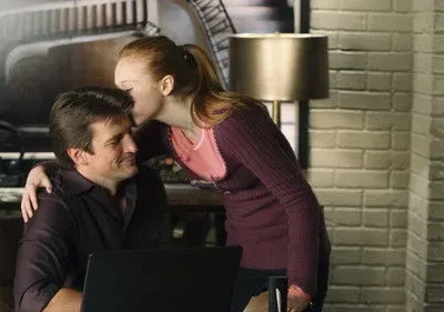Mulher ruiva beija a testa de homem sorridente sentado com laptop. Ambiente interno com parede de tijolos e luminária.