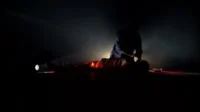 Cena escura: uma pessoa ajoelhada sobre outra deitada. Iluminada por uma lanterna e vela, o ambiente é tenso e misterioso.