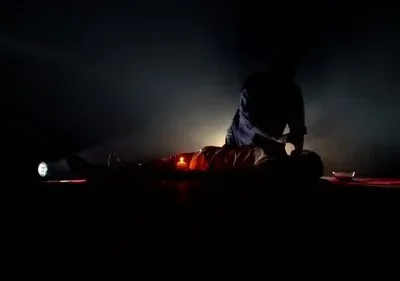 Cena escura: uma pessoa ajoelhada sobre outra deitada. Iluminada por uma lanterna e vela, o ambiente é tenso e misterioso.
