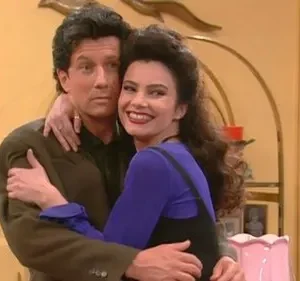 Um homem e uma mulher de cabelos escuros se abraçam. Ela sorri abertamente, ele olha para o lado. Ambiente acolhedor.