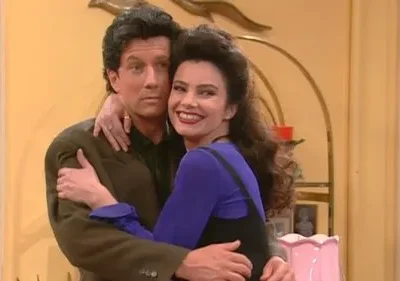 Um homem e uma mulher de cabelos escuros se abraçam. Ela sorri abertamente, ele olha para o lado. Ambiente acolhedor.