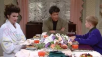 Mulher de roupão, homem e menino à mesa farta com frutas e flores. A mulher parece pensativa, enquanto os outros conversam.