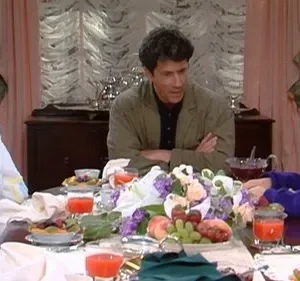 Mulher de roupão, homem e menino à mesa farta com frutas e flores. A mulher parece pensativa, enquanto os outros conversam.