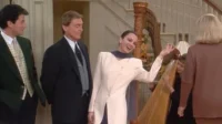 Mulher sorridente de roupa clara gesticula alegremente em salão elegante com harpa. Homens de terno e outros a observam.