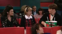 Mulher de cabelo escuro sorri entre jovem loira e rapaz com bebidas, em cinema. Assentos vermelhos e clima descontraído.