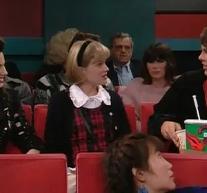 Mulher de cabelo escuro sorri entre jovem loira e rapaz com bebidas, em cinema. Assentos vermelhos e clima descontraído.