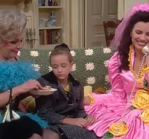 Mulher loira de boa azul alimenta criança séria. Mulher de vestido rosa volumoso com véu fala ao telefone sorrindo. Clima cômico.