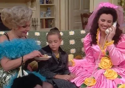 Mulher loira de boa azul alimenta criança séria. Mulher de vestido rosa volumoso com véu fala ao telefone sorrindo. Clima cômico.