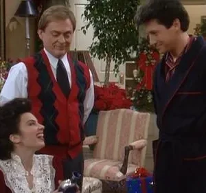 Uma mulher animada ri, sentada em sala decorada de Natal, segurando um objeto. Dois homens sorridentes a observam. Ambiente festivo.