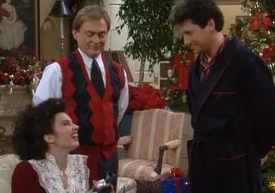 Uma mulher animada ri, sentada em sala decorada de Natal, segurando um objeto. Dois homens sorridentes a observam. Ambiente festivo.