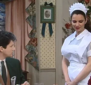 Mulher em uniforme de babá e capuz branco sorri, enquanto um homem de terno a observa numa sala de jantar formal com crianças.