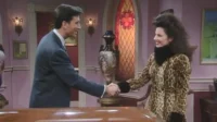 Um homem de terno e uma mulher de casaco de leopardo e cabelo volumoso se cumprimentam, sorrindo, numa sala elegante.