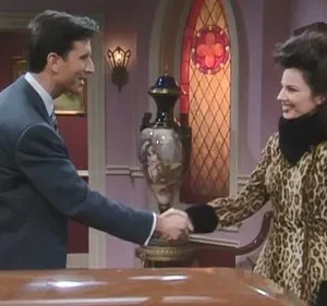 Um homem de terno e uma mulher de casaco de leopardo e cabelo volumoso se cumprimentam, sorrindo, numa sala elegante.