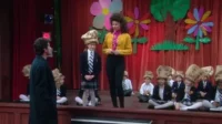 Mulher e homem observam crianças de uniforme e chapéus de saco de papel em palco decorado com flores para uma peça escolar.