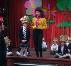 Mulher e homem observam crianças de uniforme e chapéus de saco de papel em palco decorado com flores para uma peça escolar.