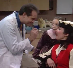 Médico sorridente, mulher em fantasia colorida e menina travessa que mostra a língua, num consultório. Clima divertido e humorístico.