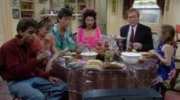 Seis pessoas, jovens e adultos, jantam numa mesa farta e casual. Uma mulher de rosa vibrante, um homem de terno. Cena doméstica animada.