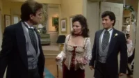 Mulher com espartilho branco/vermelho entre dois homens de terno em casa elegante. Ela parece surpresa, um homem sorri. Fundo de casamento.