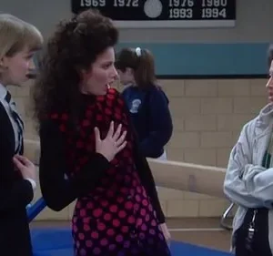 Em um ginásio escolar, três mulheres: uma de uniforme, uma morena dramática com vestido de bolinhas e uma séria de jaqueta. Cena cômica.