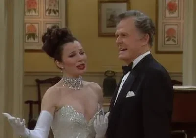 Mulher glamorosa em vestido brilhante e homem de smoking em sala luxuosa. Ela gesticula, ele sorri. Clima de evento sofisticado.