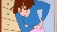 Mulher animada de cabelo castanho, camisa azul e calça rosa, curvada, com as mãos nas costas e suor na testa, indicando dor ou estresse.