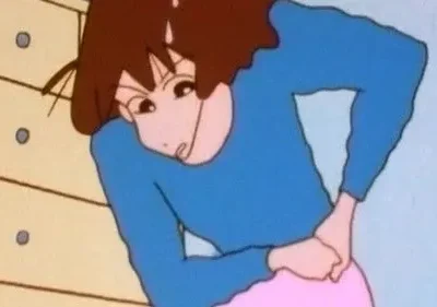 Mulher animada de cabelo castanho, camisa azul e calça rosa, curvada, com as mãos nas costas e suor na testa, indicando dor ou estresse.