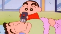 Menino de vermelho canta em microfone apontado para a barriga de um homem barbudo deitado, aparentemente dormindo. Cena cômica.