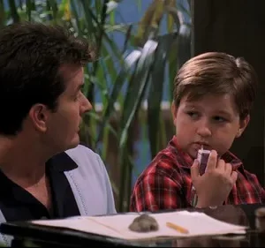 Um homem de polo casual e um menino de camisa xadrez bebem juntos. O homem olha à direita, o menino bebe de caixinha, pensativo. Fundo com plantas.