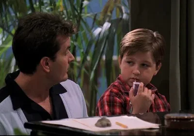 Um homem de polo casual e um menino de camisa xadrez bebem juntos. O homem olha à direita, o menino bebe de caixinha, pensativo. Fundo com plantas.