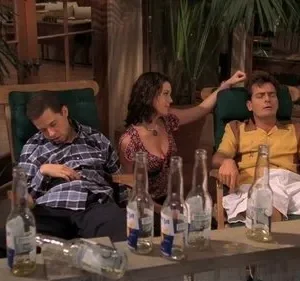 Em uma varanda, dois homens dormem exaustos em cadeiras, com uma mulher ao lado. Garrafas de cerveja vazias indicam uma festa.