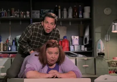 Homem surpreso inclina-se sobre mulher assustada numa lavanderia. Prateleiras bagunçadas com itens de limpeza ao fundo.